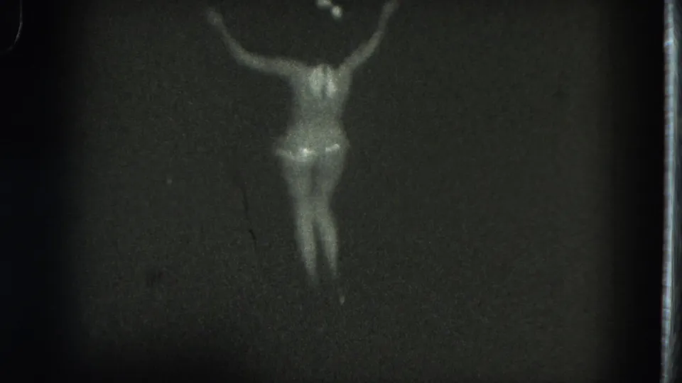 1975 San Francisco 8mm frame showing the dancer from behind with arms lifted against a dark performance backdrop.