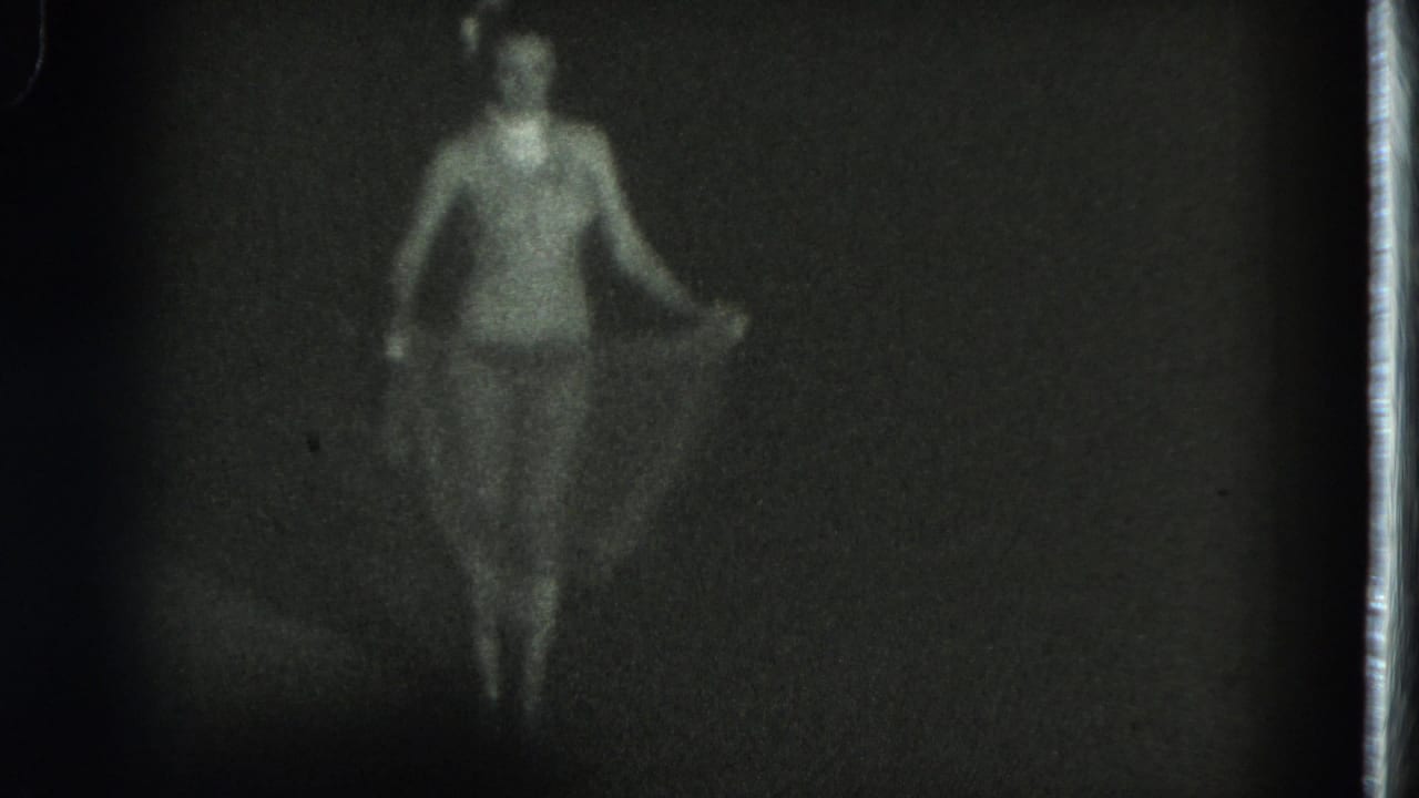 Soft 1975 8mm frame from San Francisco showing a woman in a revealing outfit standing in low light with fabric at her sides.