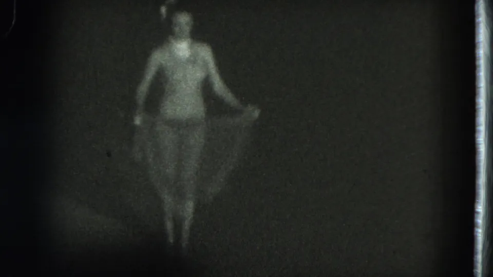 Soft 1975 8mm frame from San Francisco showing a woman in a revealing outfit standing in low light with fabric at her sides.