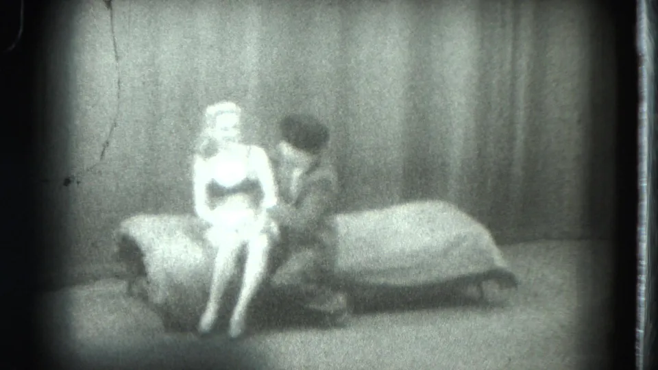 1975 8mm home-movie frame from San Francisco showing a woman seated on a bed as another adult leans close in a curtained interior.