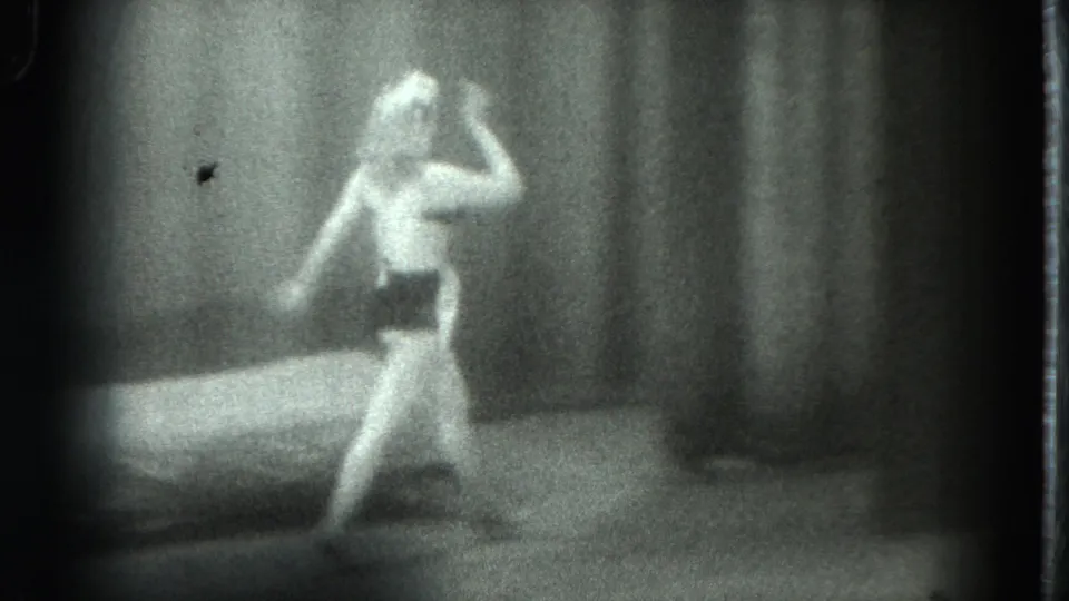 1975 San Francisco 8mm frame showing a woman in a revealing outfit moving beside a bed against a curtain backdrop.