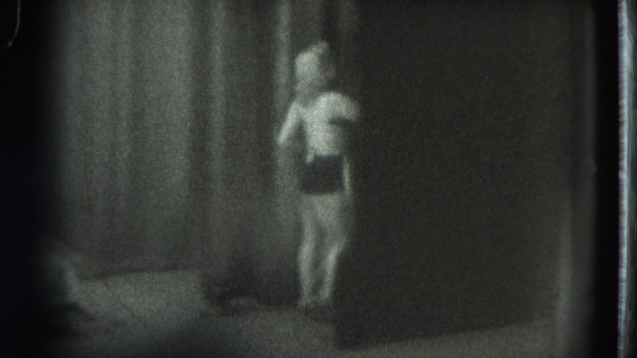1975 8mm frame from San Francisco showing a woman walking through a dim private room with a bed nearby.