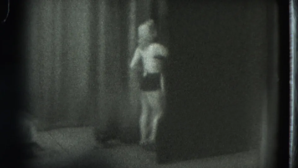 1975 8mm frame from San Francisco showing a woman walking through a dim private room with a bed nearby.