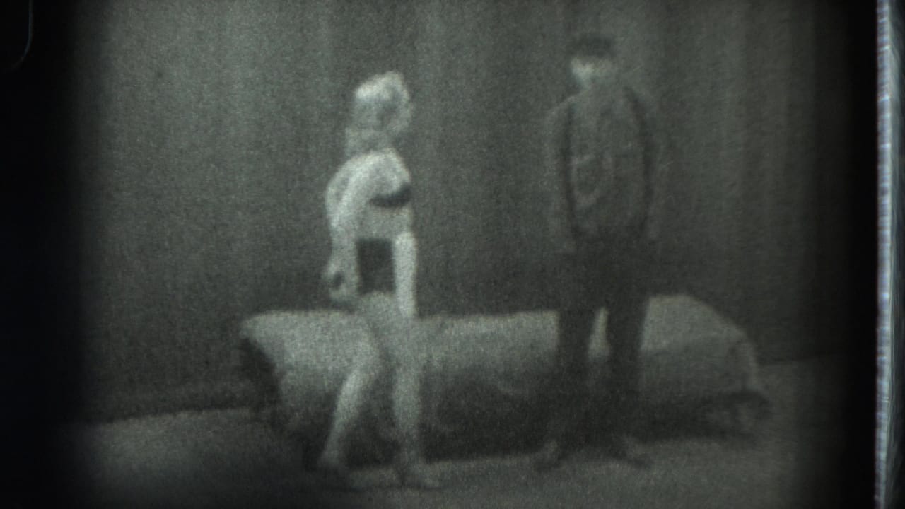 Woman near a bed with a man standing farther back in a dim interior.