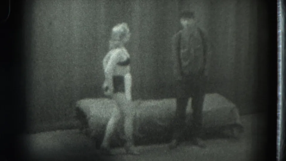 1975 San Francisco home-movie frame with a woman near a bed and a man standing in the background of a private room.