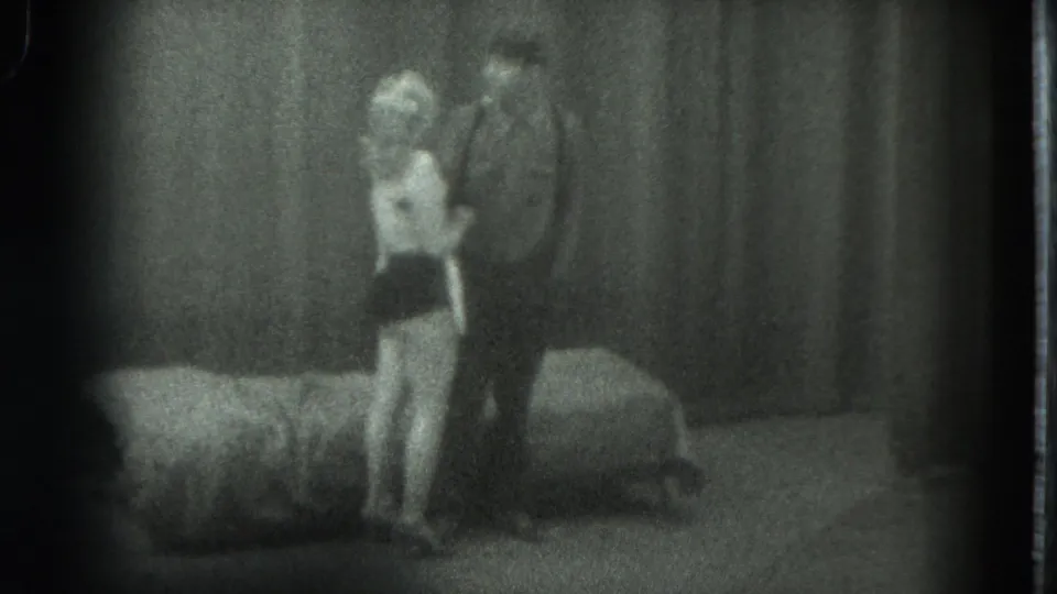 Grainy 1975 8mm frame from San Francisco showing two adults standing near a bed in a private interior.