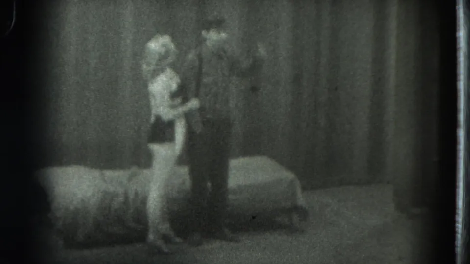 1975 San Francisco 8mm frame showing a woman and man facing each other near a bed in a private room.