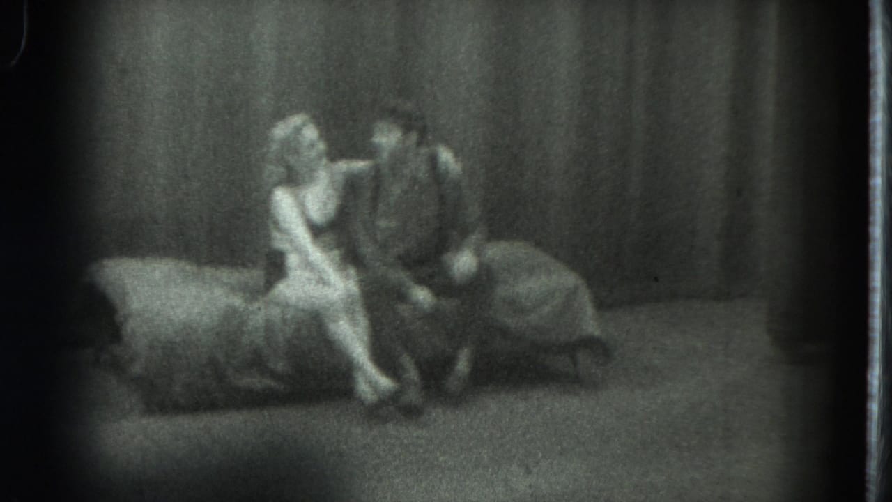 1975 San Francisco 8mm frame showing a woman in light clothing seated close to a man on a couch before heavy curtains.