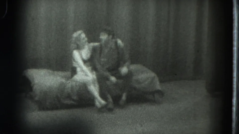 1975 San Francisco 8mm frame showing a woman in light clothing seated close to a man on a couch before heavy curtains.