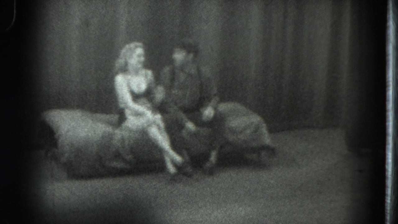Woman seated with crossed legs beside a man on a couch in front of curtains.