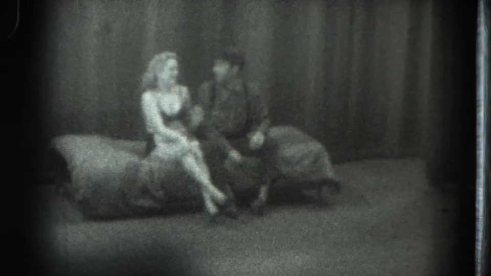 1975 San Francisco home-movie frame with a woman sitting beside a man on a low couch in a dim private interior.