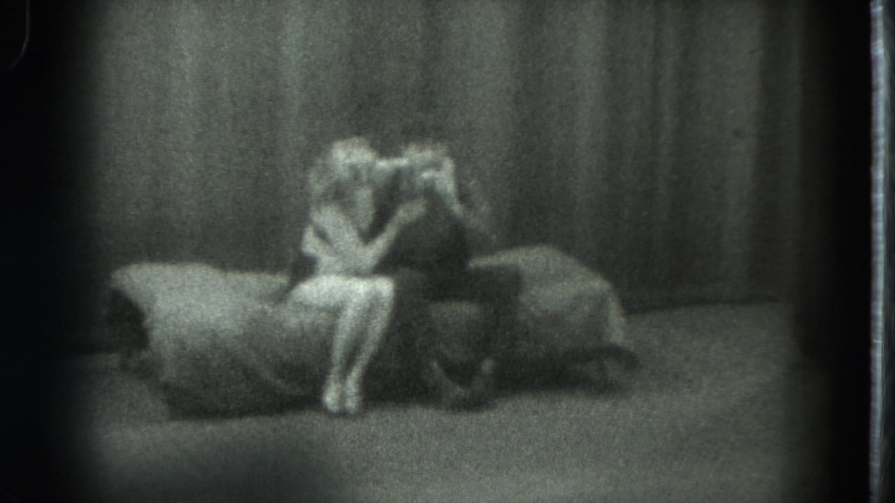 1975 San Francisco 8mm frame with two stage performers leaning together near a bed prop in low light.