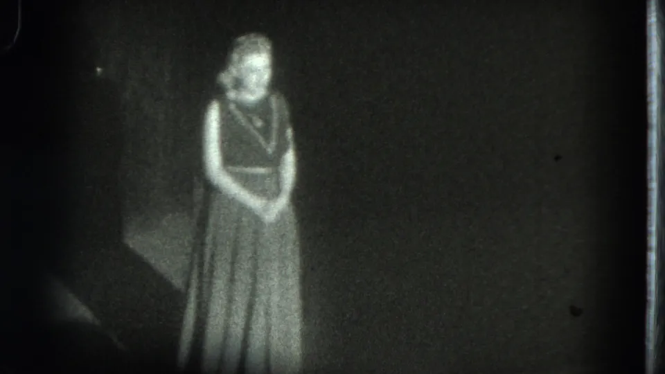 1975 San Francisco 8mm frame showing a woman in a long dress standing alone in a dark stage setting.