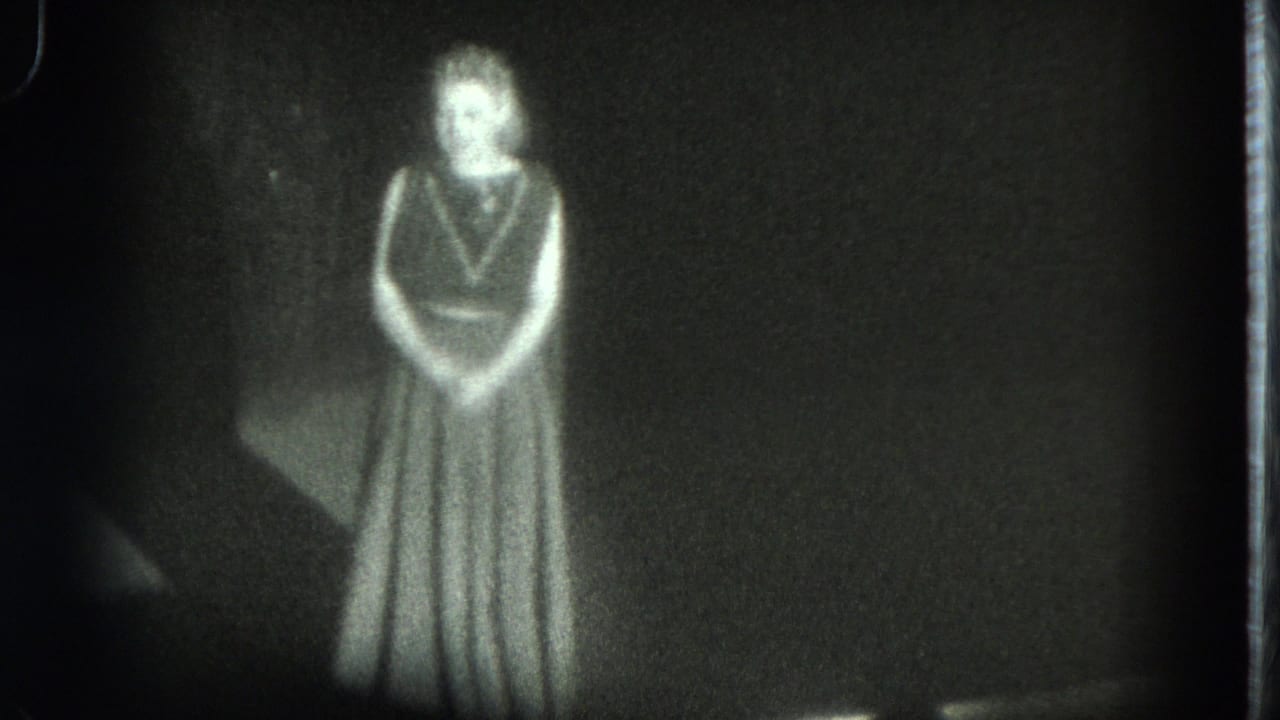 1975 San Francisco 8mm frame showing a woman in a long dress facing the audience on a dark stage.