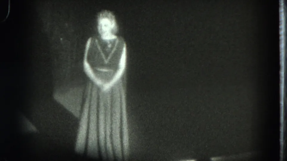 1975 San Francisco 8mm frame showing a woman in a long dress facing the audience on a dark stage.