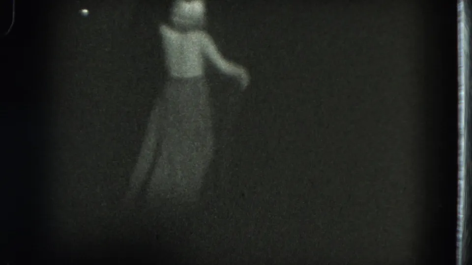 A performer moves across the dim stage in a long skirt, captured in soft, grainy 1975 San Francisco 8mm footage.