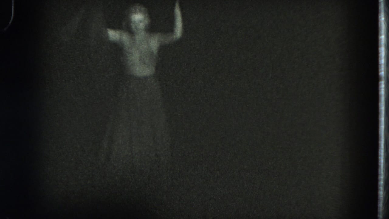 In this 1975 San Francisco 8mm frame, a woman appears in low light with one arm lifted against a nearly black background.