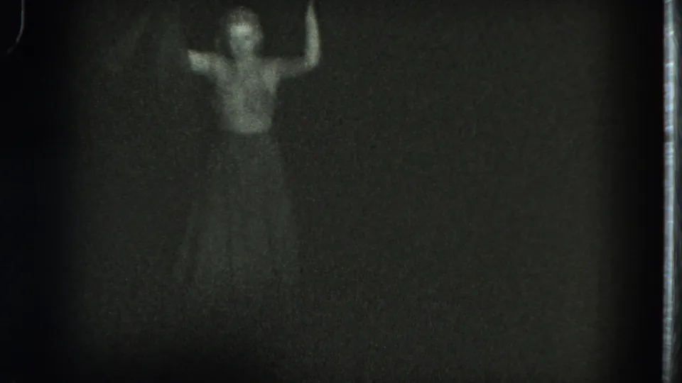 In this 1975 San Francisco 8mm frame, a woman appears in low light with one arm lifted against a nearly black background.
