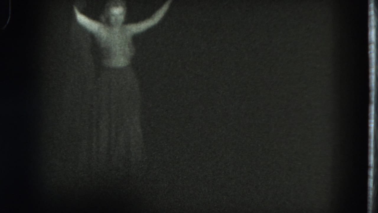 Woman faces the camera with both arms lifted in a dark, grainy room.