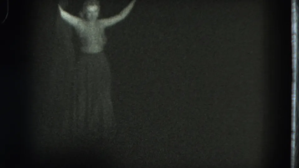 A low-light 8mm frame shows the woman facing forward with both arms raised, her body softly rendered by grain and exposure.