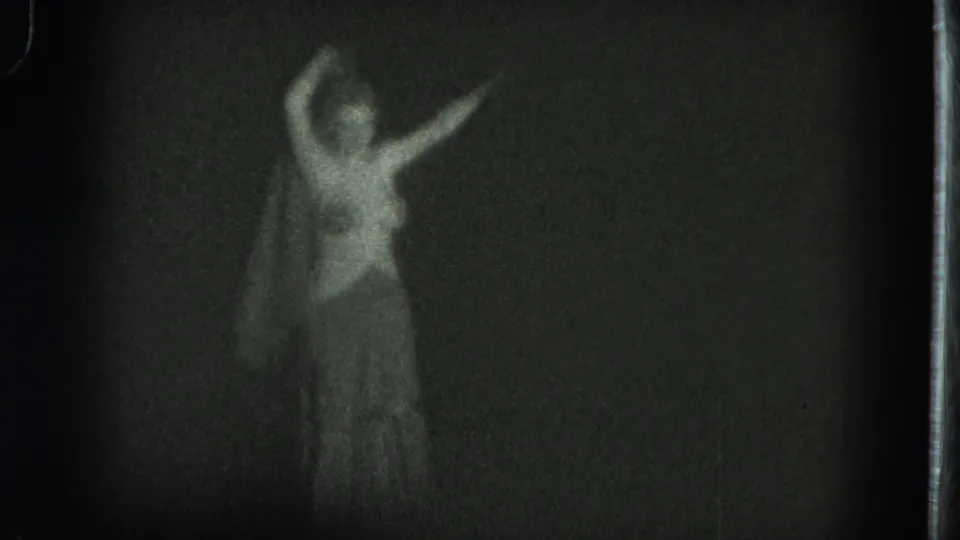 The woman appears mid-turn in the 1975 8mm footage, one arm above her head and another extended through the dim interior light.