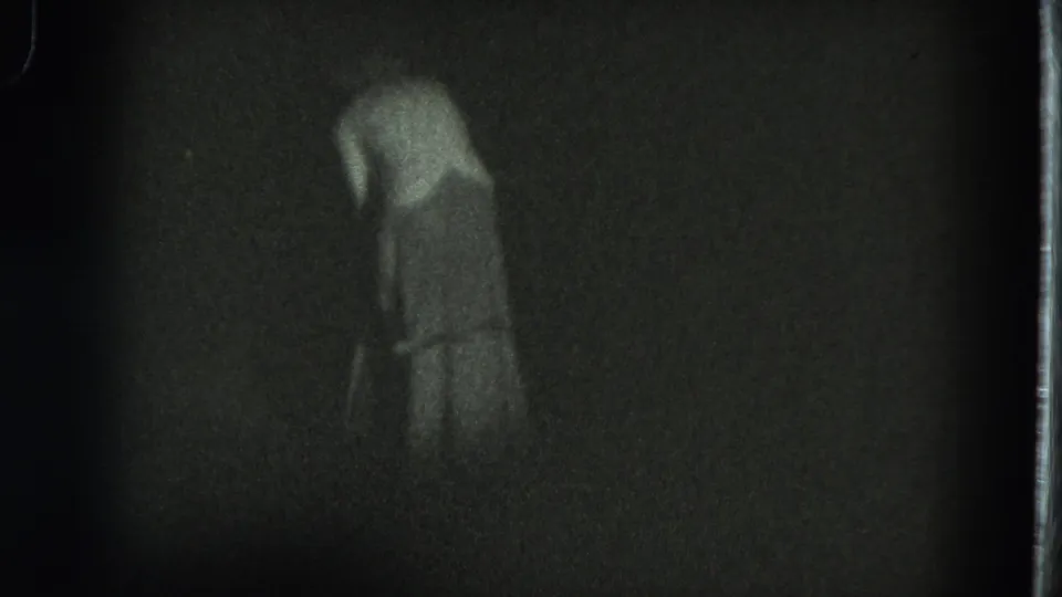 Near the end of the clip, the woman bends forward in the dim 8mm frame, with her figure softened by grain and shadow.