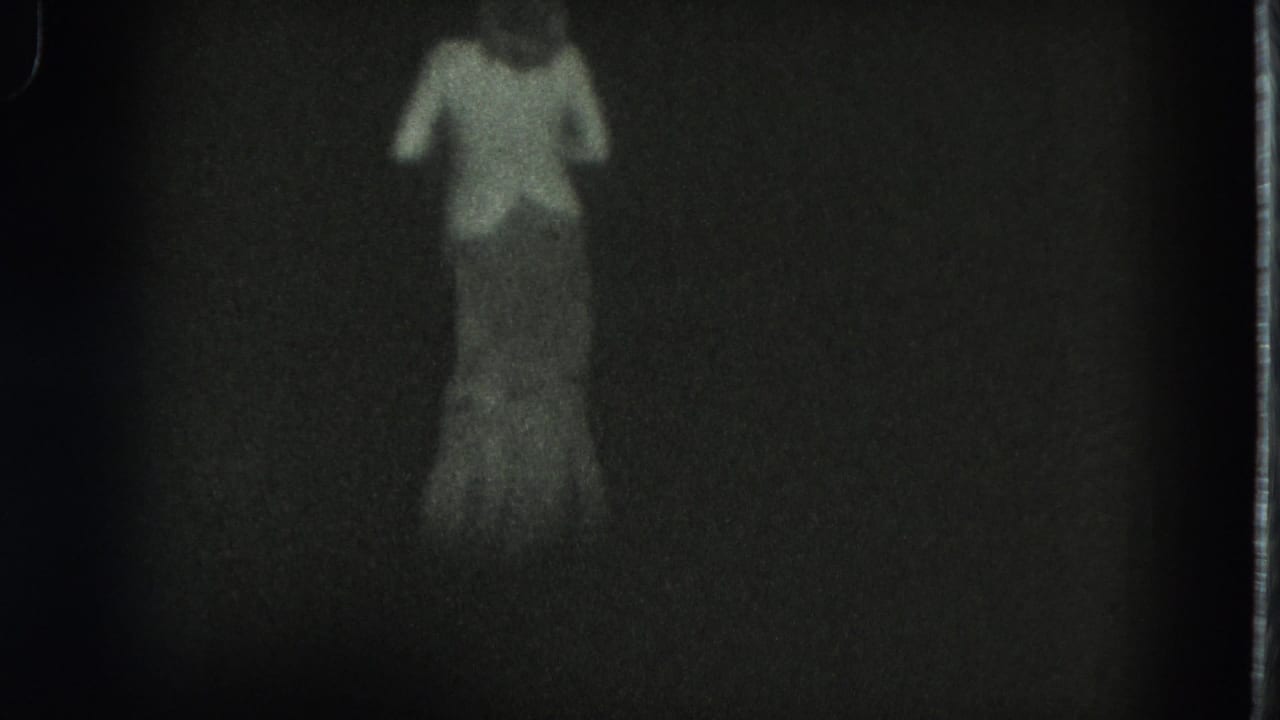 Low-light 8mm frame from San Francisco in 1975 with a woman standing or walking as her long dress blends into the dark scene.