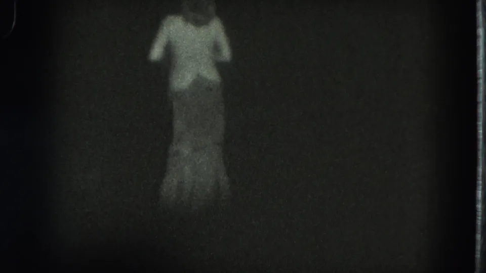 Low-light 8mm frame from San Francisco in 1975 with a woman standing or walking as her long dress blends into the dark scene.