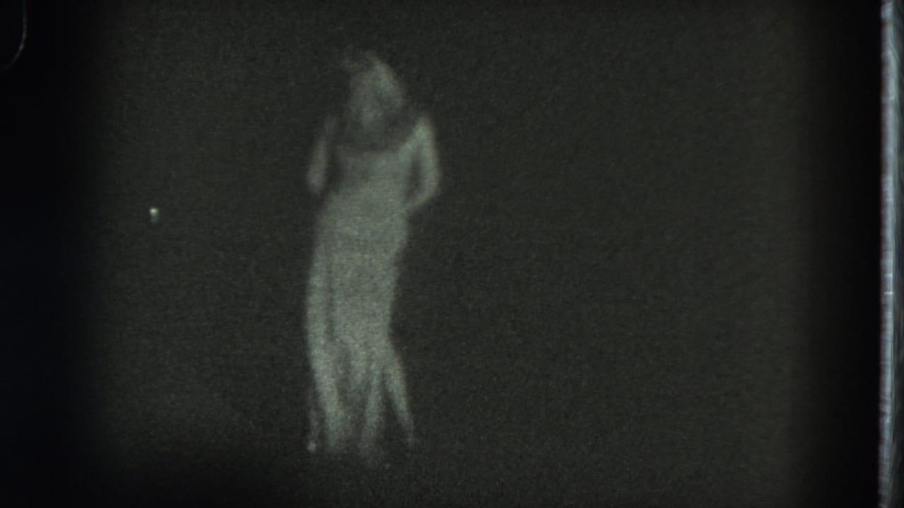 The dancer turns in a long white dress under dim light, with the 8mm frame soft and grainy.