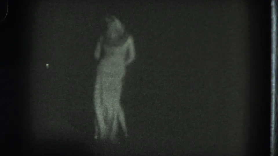 The dancer turns in a long white dress under dim light, with the 8mm frame soft and grainy.