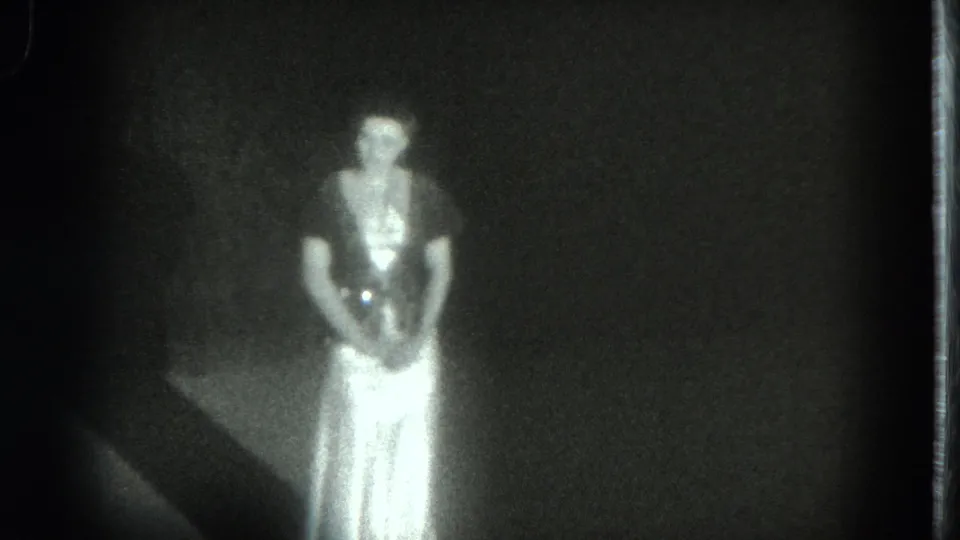 The dancer remains facing forward in the dim scene, with a dark floor edge visible beside her.