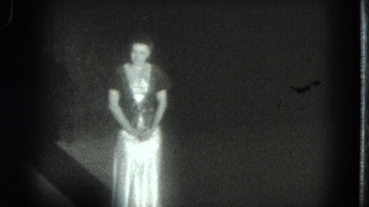 In 1975 San Francisco home-movie footage, a woman in a pale flowing dress faces the camera in a dark interior.