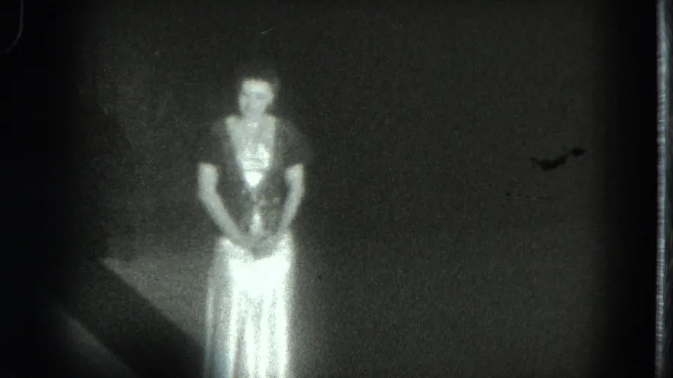 In 1975 San Francisco home-movie footage, a woman in a pale flowing dress faces the camera in a dark interior.