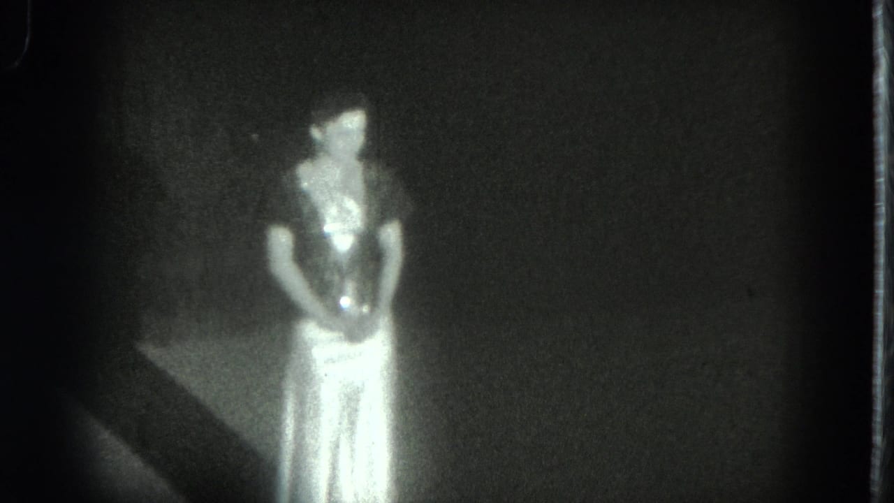 The woman remains facing the camera in a dim 1975 San Francisco interior, with her pale dress catching most of the light.
