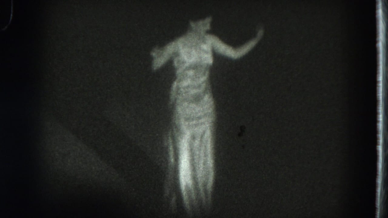 1975 San Francisco 8mm frame of a dancer in a long white dress, softly lit against a dark stage backdrop.