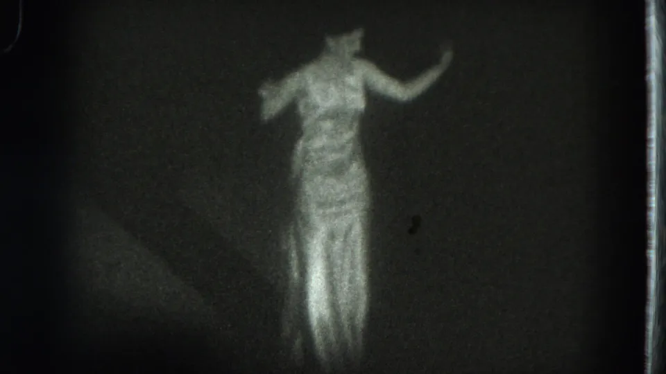 1975 San Francisco 8mm frame of a dancer in a long white dress, softly lit against a dark stage backdrop.