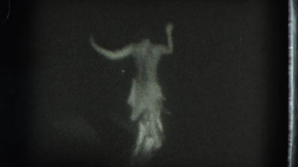 The performer appears in motion with arms extended, the white costume glowing softly in the dark 8mm stage frame.