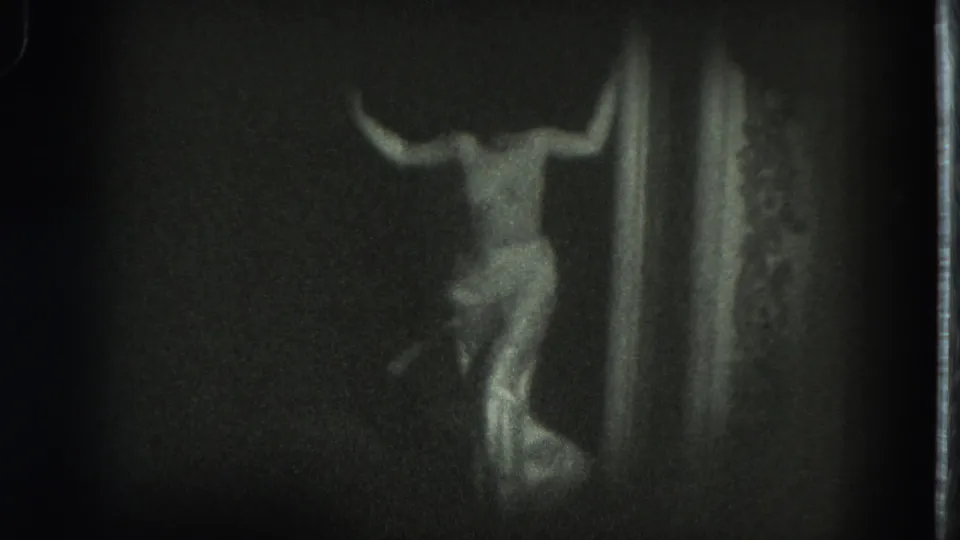 A dancer in a light costume appears near a vertical stage element, captured from an audience viewpoint on grainy 8mm film.