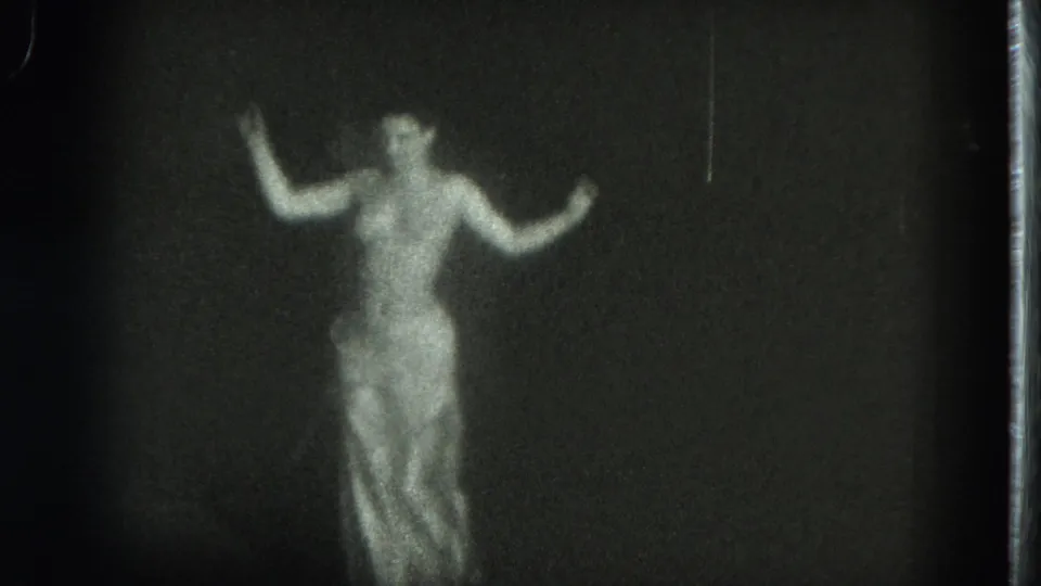 The white-dressed performer faces the camera with both arms lifted, isolated against a black stage background.