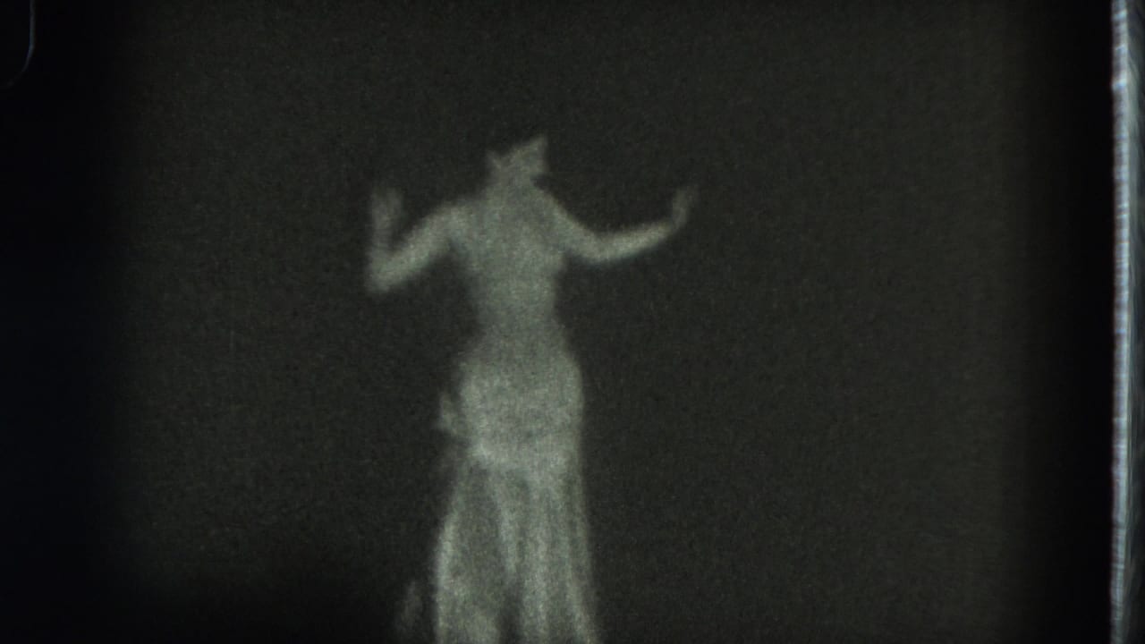 Woman in a shiny gown dances against a dark background with both arms raised.