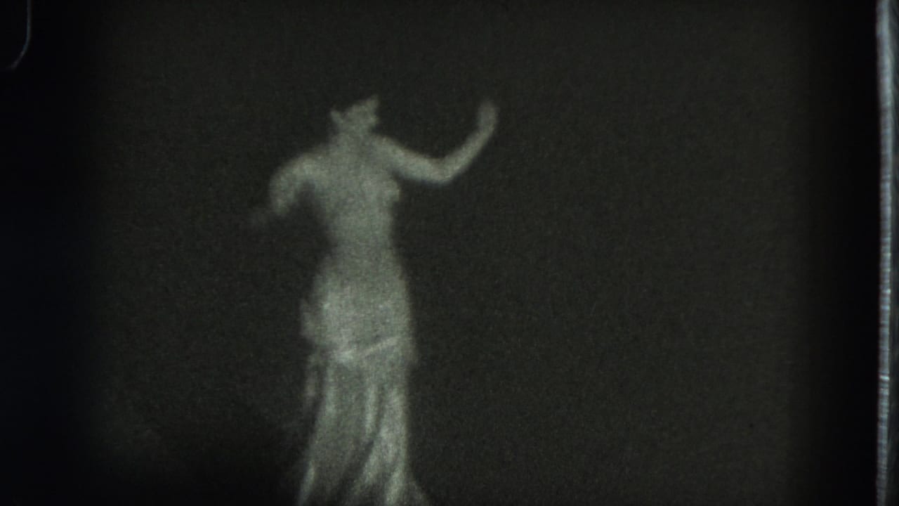 The dancer remains centered in the grainy 8mm frame, her reflective gown catching light as one arm curves upward.