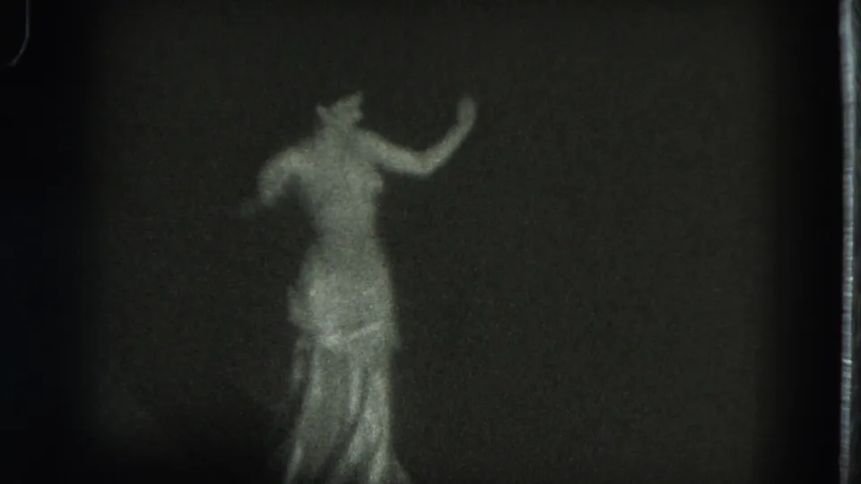 The dancer remains centered in the grainy 8mm frame, her reflective gown catching light as one arm curves upward.