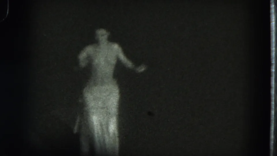 In 1975 San Francisco 8mm footage, an adult dancer in a flowing skirt raises one arm while facing away in soft focus.