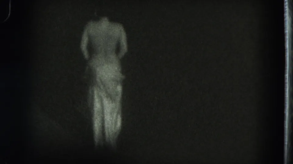 A rear view of the adult dancer shows a lowered upper garment or bare shoulders above a light skirt in grainy 8mm footage.