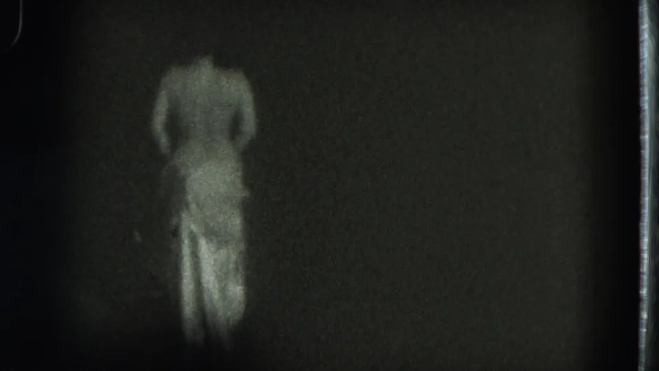 The adult dancer moves in soft focus, with a light skirt and partially lowered dress visible against the dark 8mm frame.