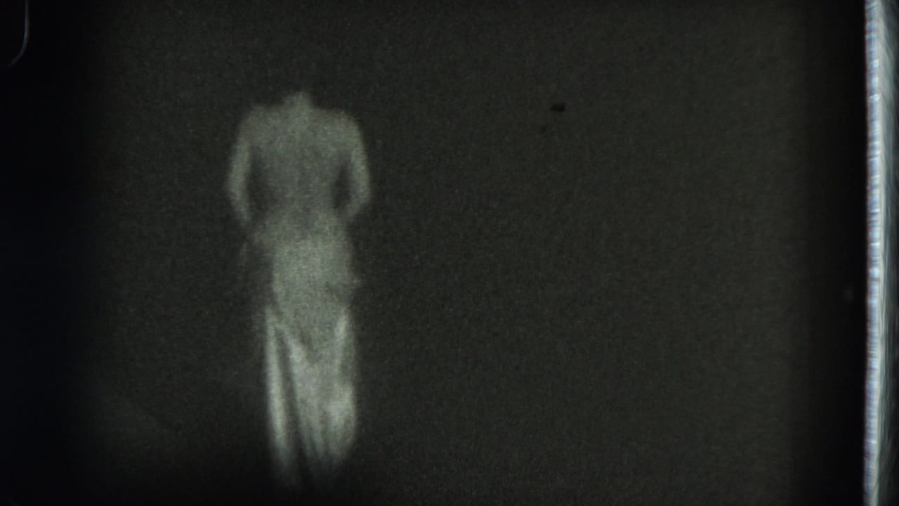 1975 8mm frame from San Francisco metadata showing a topless adult woman from behind in a light skirt against a dark background.