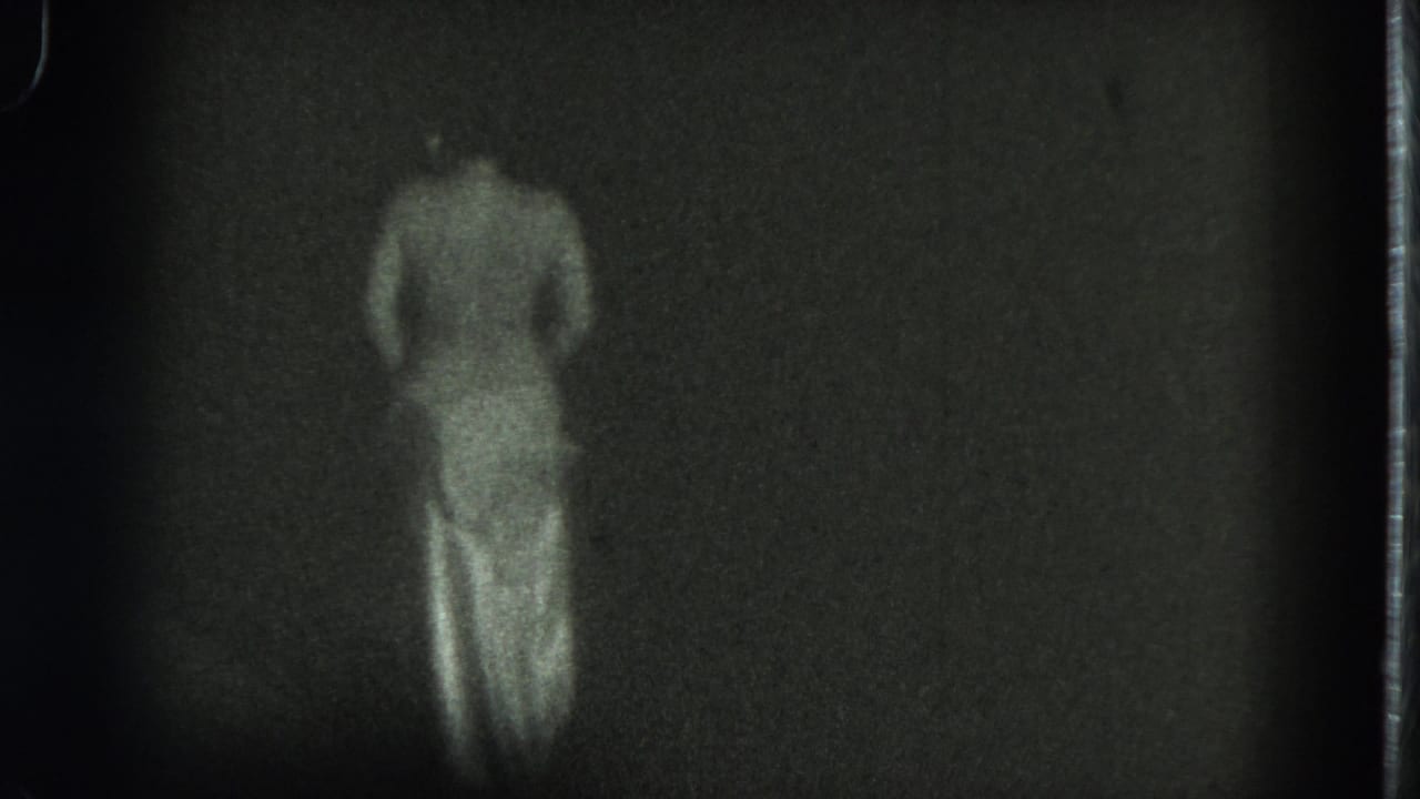 1975 San Francisco 8mm frame showing a topless adult woman from behind as she shifts position in a dark, undefined space.