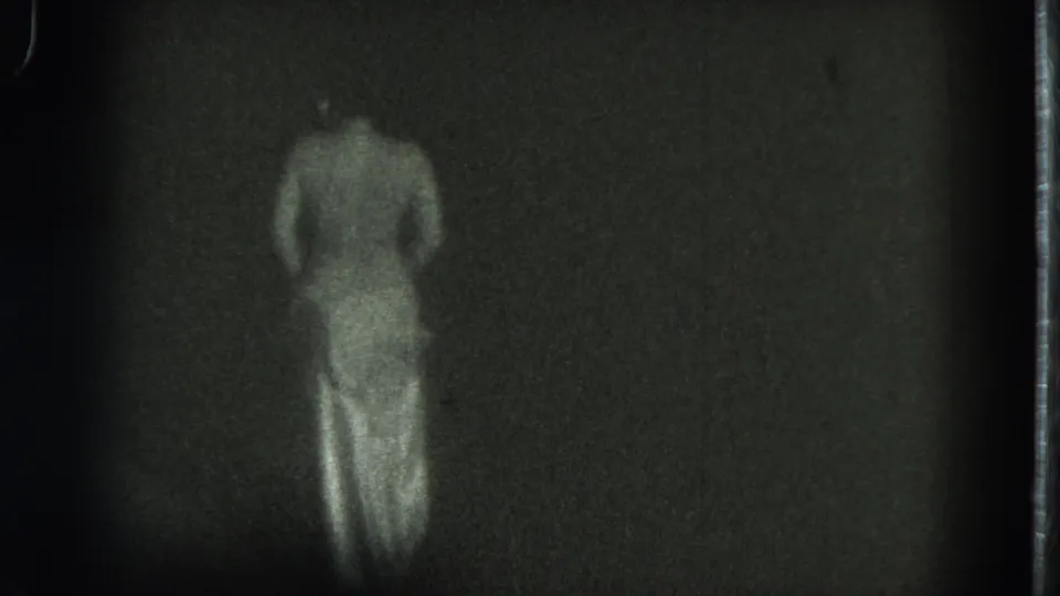 1975 San Francisco 8mm frame showing a topless adult woman from behind as she shifts position in a dark, undefined space.