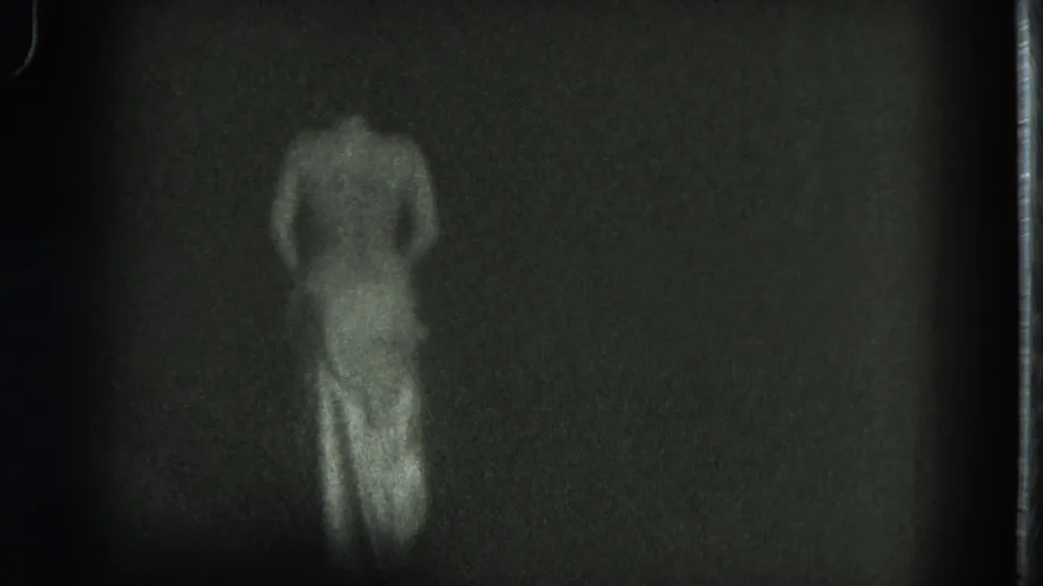 1975 8mm frame with a topless adult woman from behind, her body lower in frame as she sways in a dark setting.