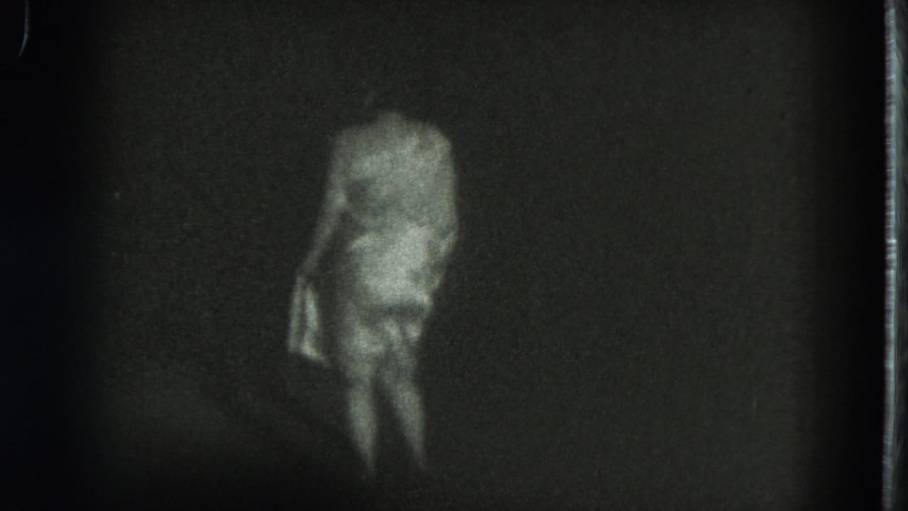 Woman in a light dress standing turned slightly away in a dark, grainy frame.